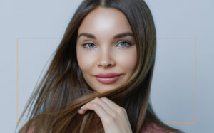 Portrait of a smiling woman with a hair style, touching her face, against a pale background. - Scott J Salons in New York, NY