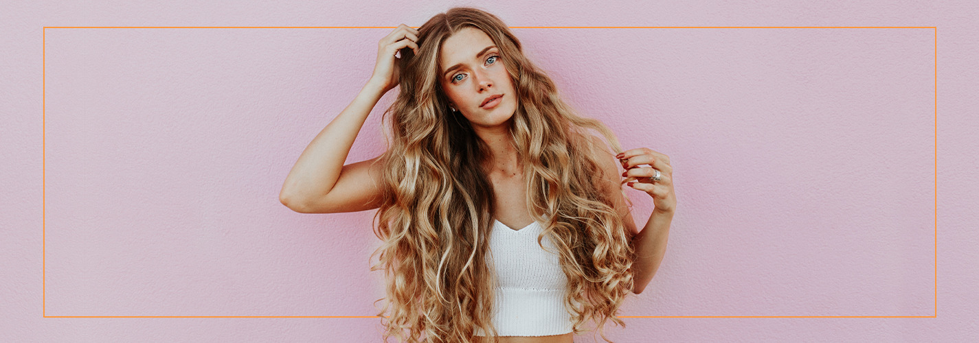 A woman with long wavy hair extensions posing against a pink background. - Scott J Salons in New York, NY