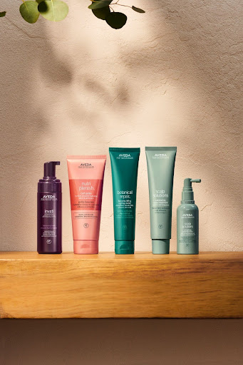 Five Aveda haircare and hair treatments products arranged in a row on a wooden shelf against a textured wall, with a plant shadow overhead. - Scott J Salons in New York, NY