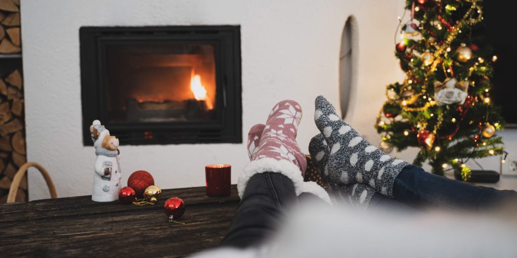 Cozy winter scene with two people relaxing by a fireplace with their feet up, wearing warm socks, near a decorated christmas tree. - Scott J Salons in New York, NY