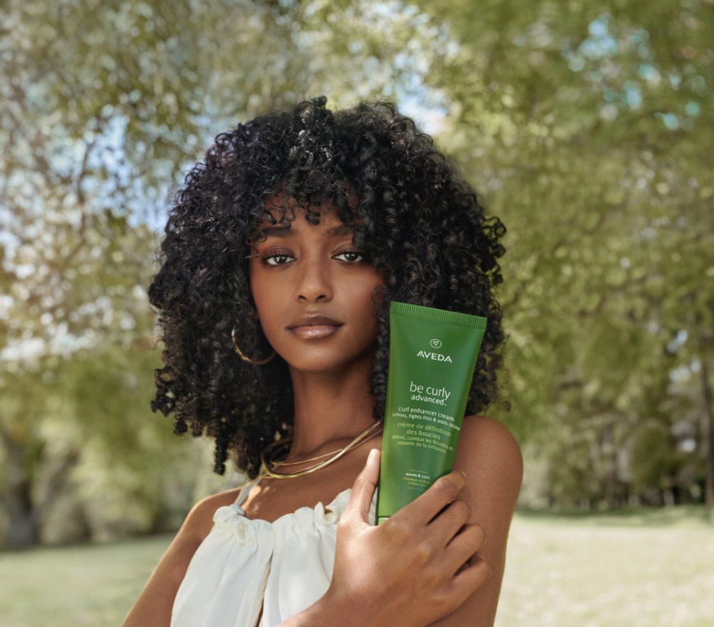 A woman with curly hair holding a bottle of aveda be curly product in a park setting. - Scott J Salons in New York, NY