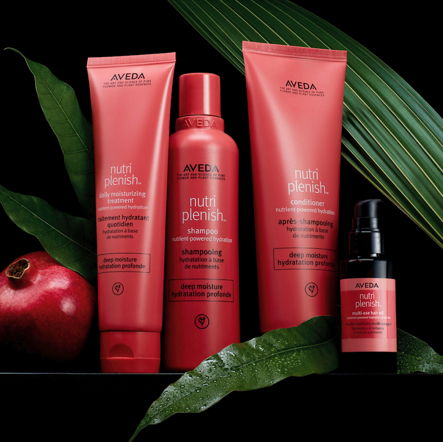 A collection of Aveda Nutriplenish hair care products, including shampoo, conditioner, and a bottle of oil, arranged with green leaves and a pomegranate in the background. - Scott J Salons in New York, NY