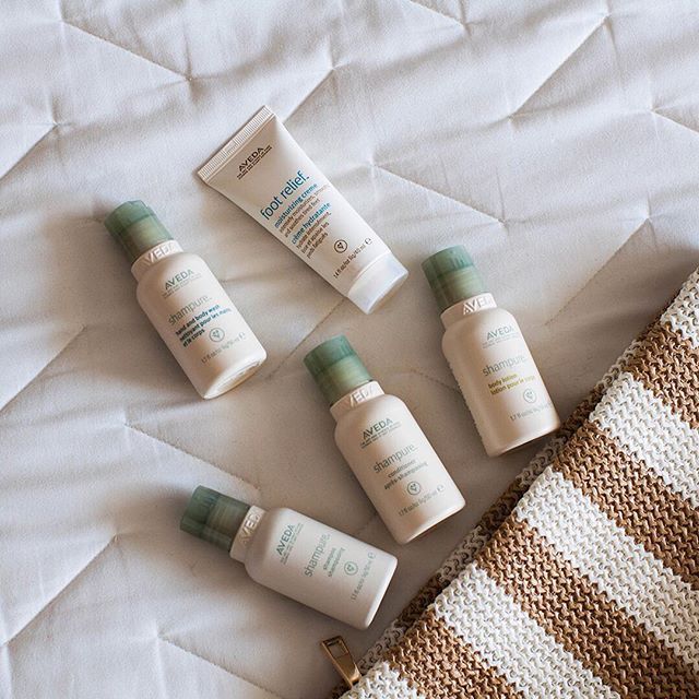 Five small bottles of Aveda hair and skin products are arranged on a white quilted surface next to a striped fabric bag. - Scott J Salons in New York, NY