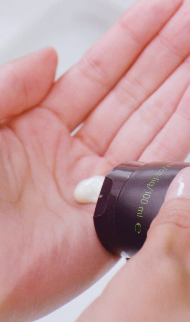 A person dispensing white cream from a dark bottle onto their palm. - Scott J Salons in New York, NY