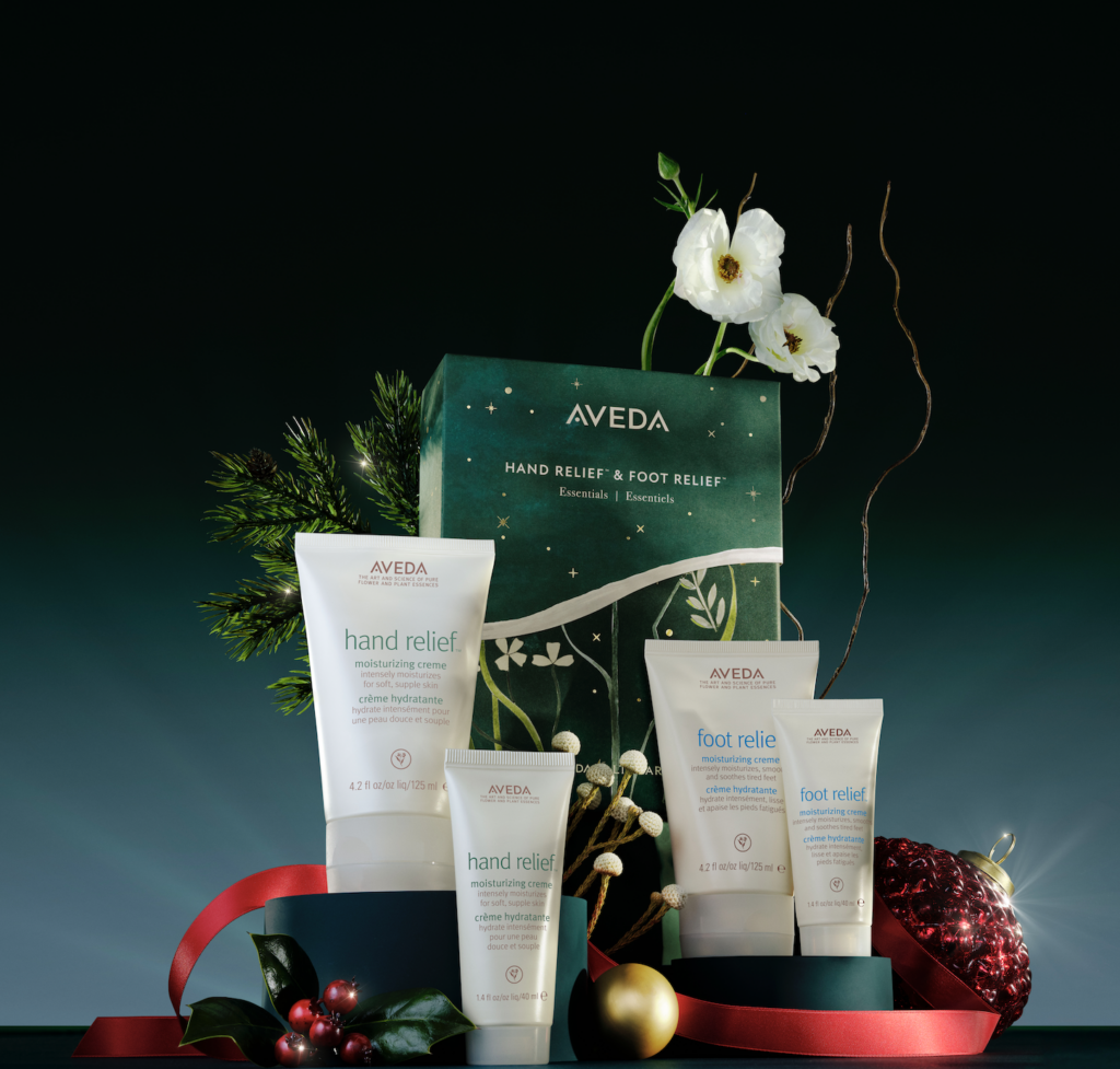 A gift set featuring Aveda products, including hand and foot creams, is displayed with decorative flowers, greenery, and ornaments on a dark background. - Scott J Salons in New York, NY