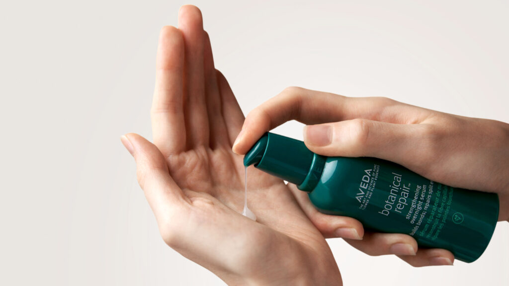 Person dispensing Aveda botanical repair serum into their hand from a green bottle. - Scott J Salons in New York, NY