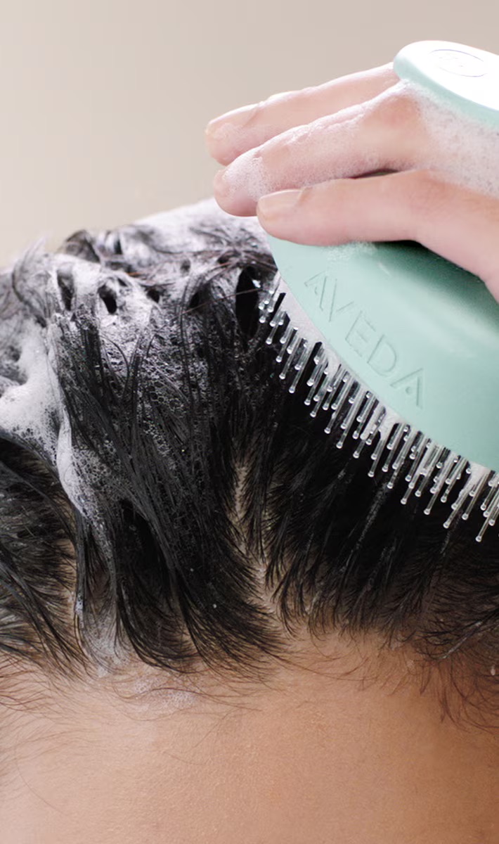 A person uses a light blue scalp massager to lather shampoo into their wet, dark hair. - Scott J Salons in New York, NY