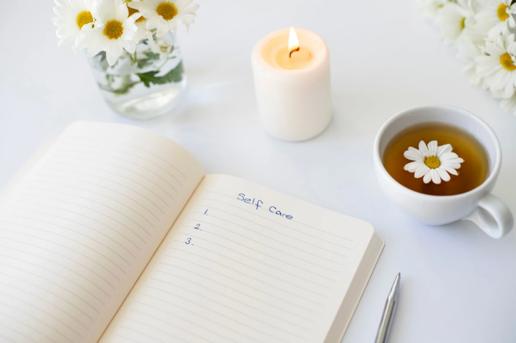 Open notebook listing "Self Care," a pen, lit candle, cup of tea with a daisy, and vase of white daisies on a table. - Scott J Salons in New York, NY