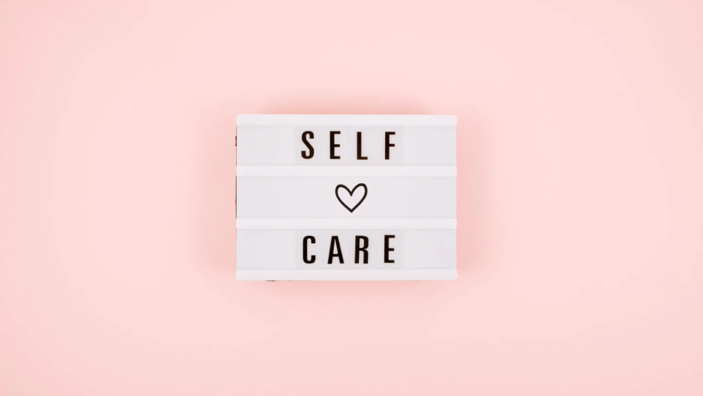 Lightbox sign with "SELF CARE" written in black letters and a heart symbol in between on a pink background. - Scott J Salons in New York, NY