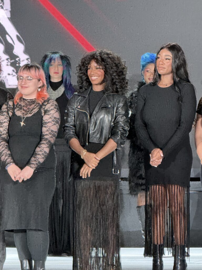 A group of women wearing black dresses stand in a row on stage, some with colored hair, in front of a dark background with red and white accents. - Scott J Salons in New York, NY