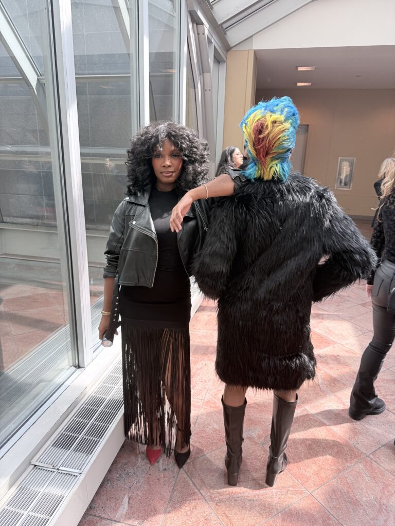 Two people pose indoors; one wears a black fringe dress and leather jacket, while the other faces away, wearing a black fur coat and has short, multicolored hair. - Scott J Salons in New York, NY