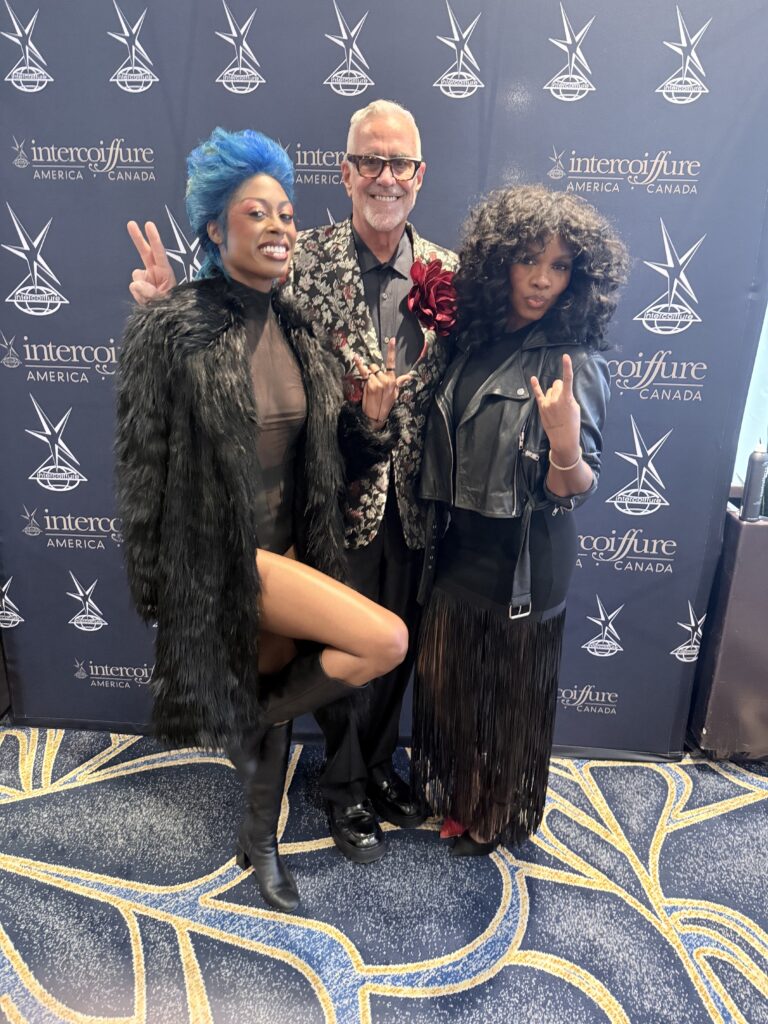 Three people posing together at an Intercoiffure America Canada event, standing in front of a branded backdrop, dressed in stylish, bold outfits. - Scott J Salons in New York, NY