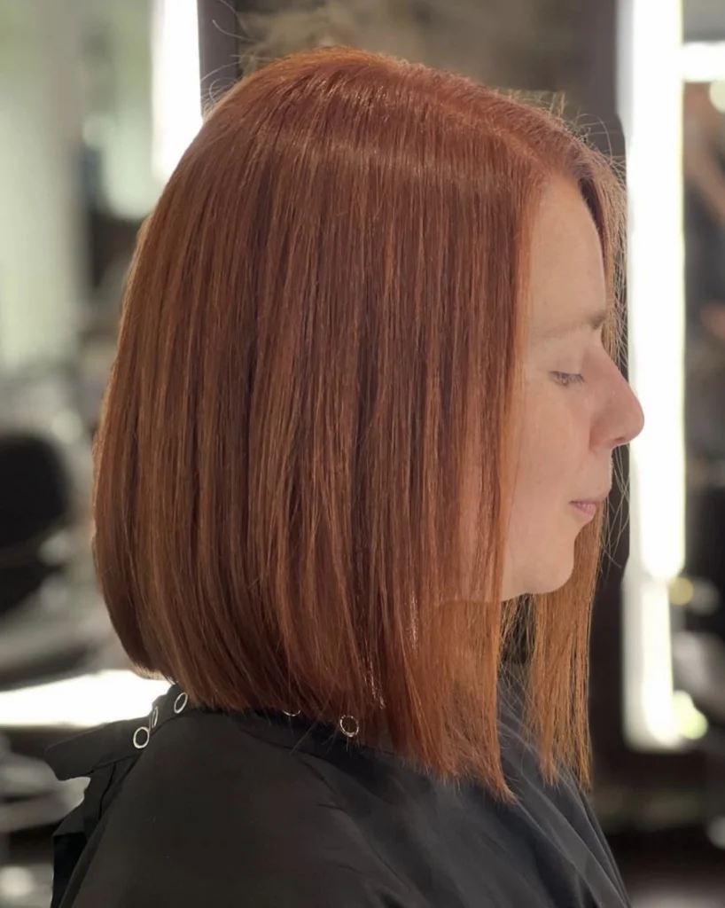A woman with straight, chin-length auburn hair sits in a salon chair, wearing a black cape, facing sideways. - Scott J Salons in New York, NY