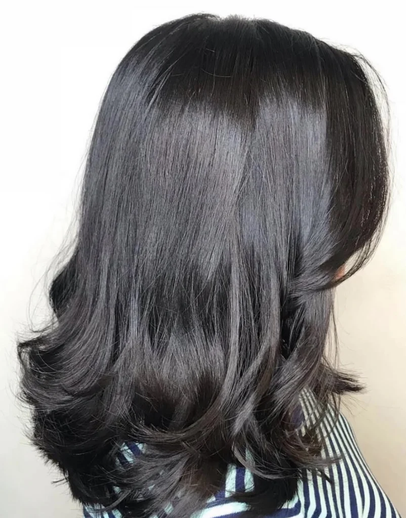 A person with shoulder-length, straight dark hair featuring soft waves at the ends, wearing a striped shirt, facing away from the camera. - Scott J Salons in New York, NY