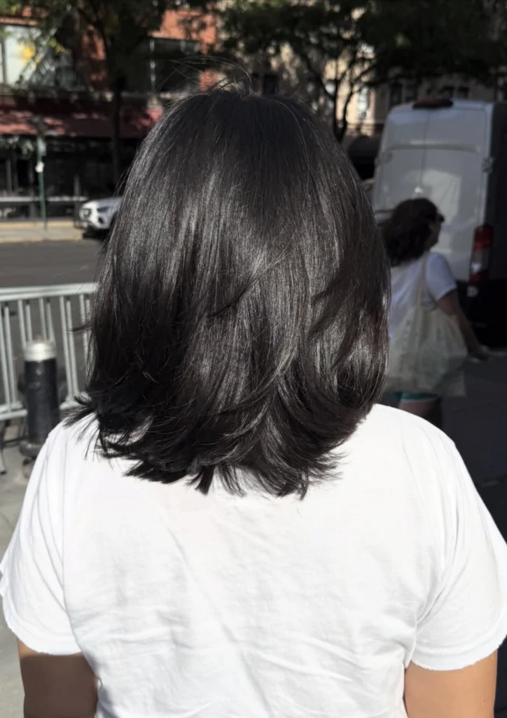 Person with shoulder-length dark hair wearing a white T-shirt stands outdoors with their back to the camera, on a city street. - Scott J Salons in New York, NY