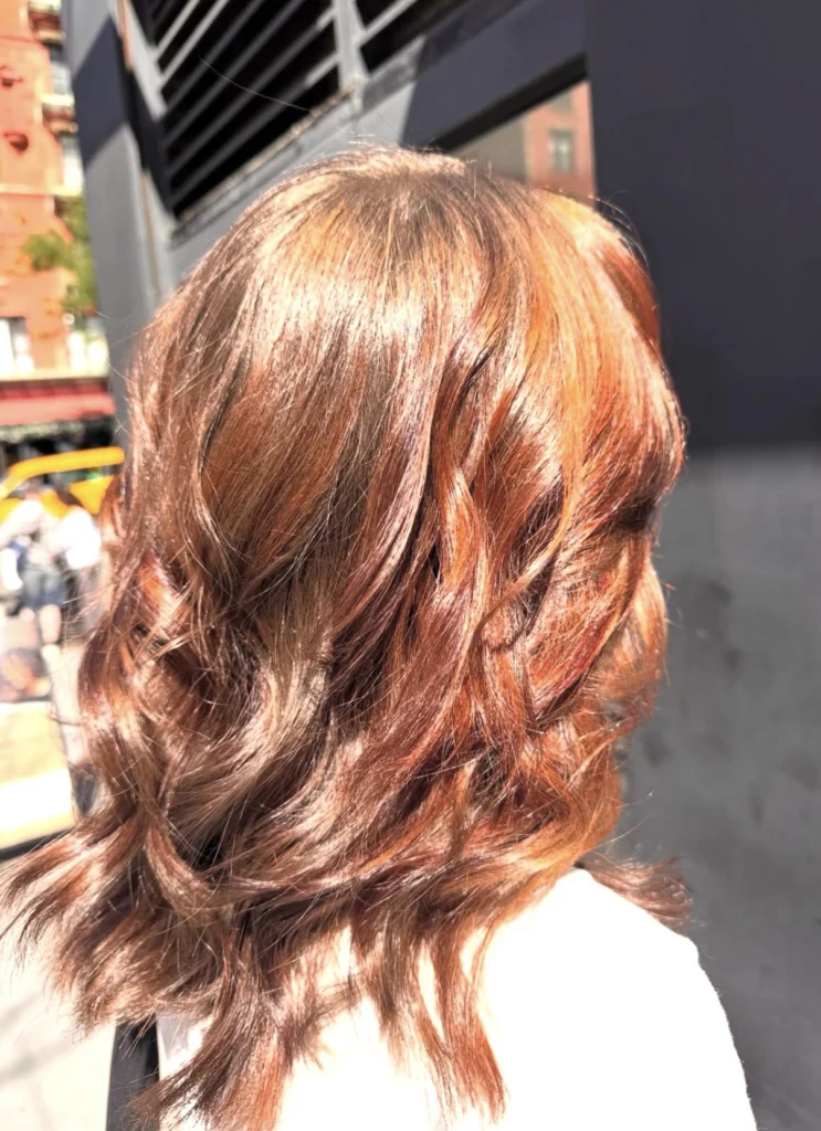A person with medium-length, wavy, brown hair with copper highlights, standing outdoors in bright sunlight. - Scott J Salons in New York, NY