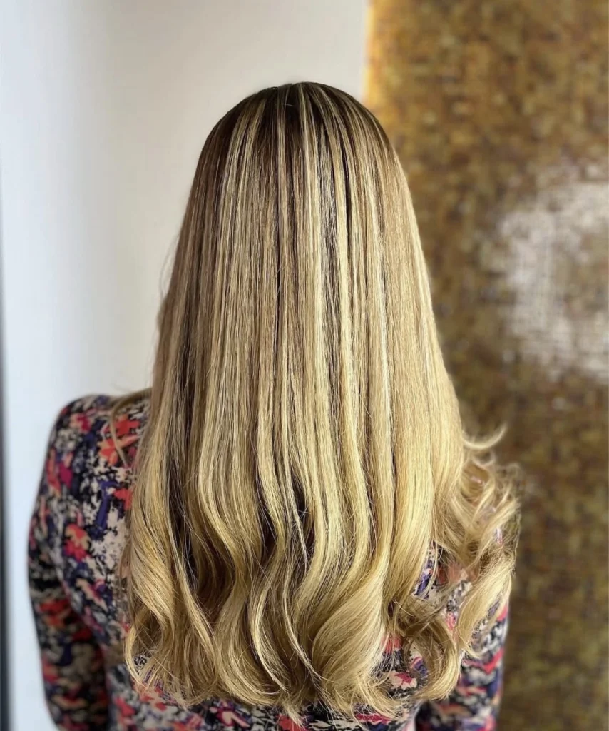 A person with long, straight, blonde hair with loose waves at the ends, wearing a patterned top, is shown from the back. - Scott J Salons in New York, NY