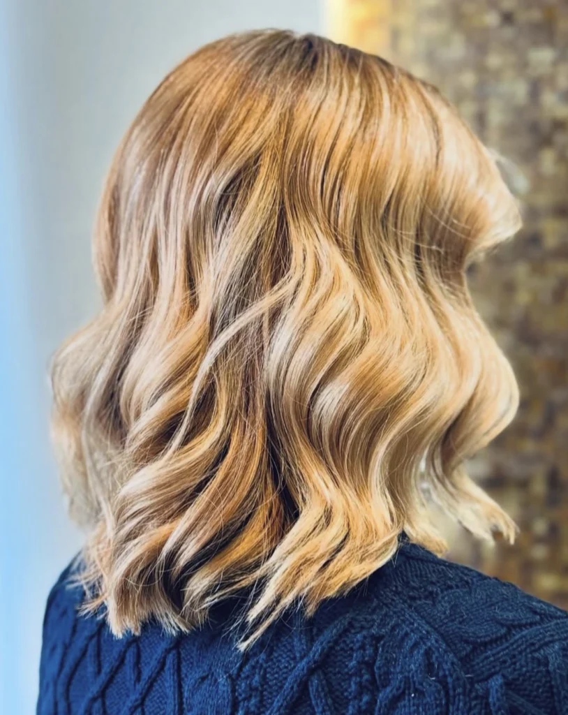 A person with shoulder-length, wavy, blonde hair, wearing a dark blue textured sweater, is shown from the back against a blurred indoor background. - Scott J Salons in New York, NY
