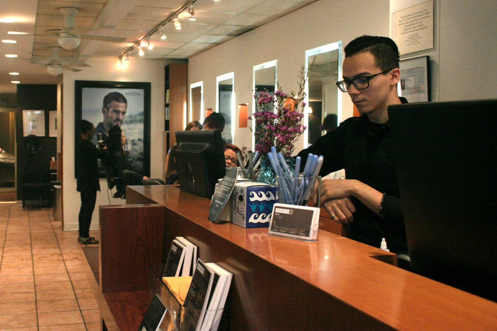 A person stands behind a salon reception desk, checking something, while other people wait or converse in the background. The desk holds pens, brochures, and a small flower arrangement. - Scott J Salons in New York, NY