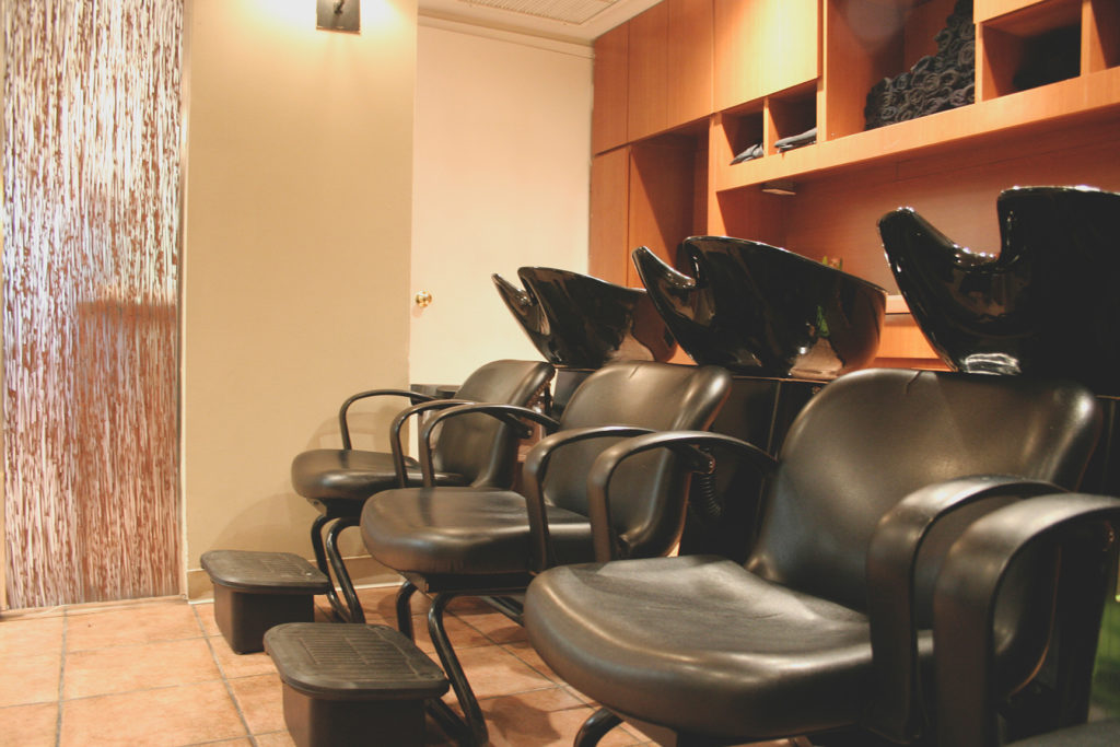 A row of black salon chairs with wash basins in a hair salon, set against shelves and a partition wall. - Scott J Salons in New York, NY