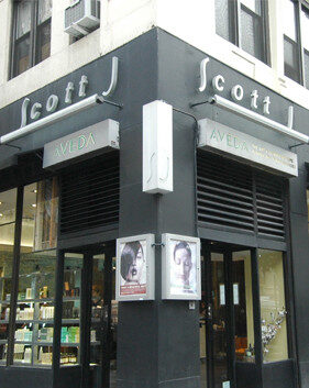 Street view of the corner entrance to Scott J Aveda Salon, featuring large windows displaying hair and beauty products. - Scott J Salons in New York, NY
