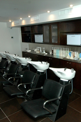 Four black salon wash chairs with white sinks are lined up in front of dark cabinets and shelves holding bottles and hair products in a modern, well-lit salon. - Scott J Salons in New York, NY