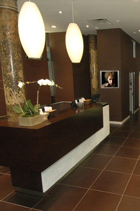 Hotel reception desk with a white orchid arrangement, pendant lights, and a wall-mounted TV displaying a person's face. The space features brown walls and tiled flooring. - Scott J Salons in New York, NY